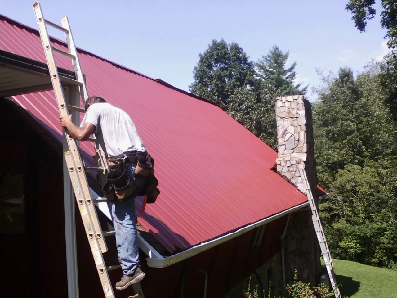 Roofer on a ladder beside a red metal roof and stone chimney — Soffit & Fascia Repair by Roofing Brothers in Agawam